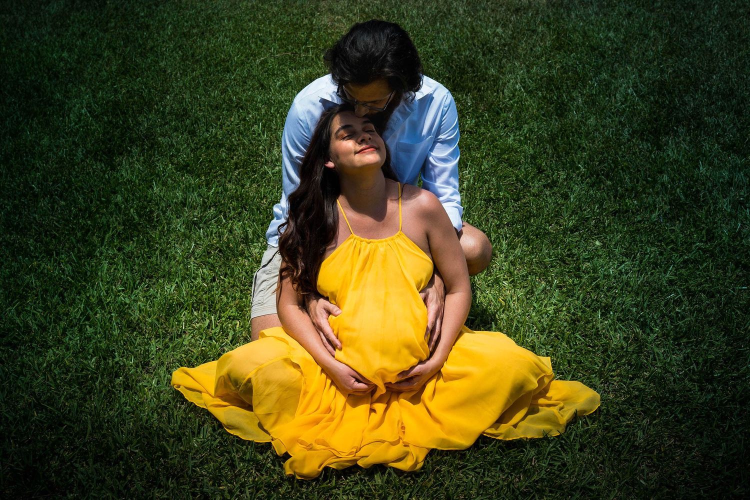 Expecting mother sitting in a sunlit garden while her partner kisses the top of her head