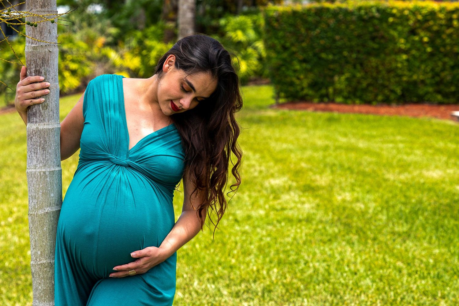 Expecting mother in a teal dress leaning against a tree trunk in a garden