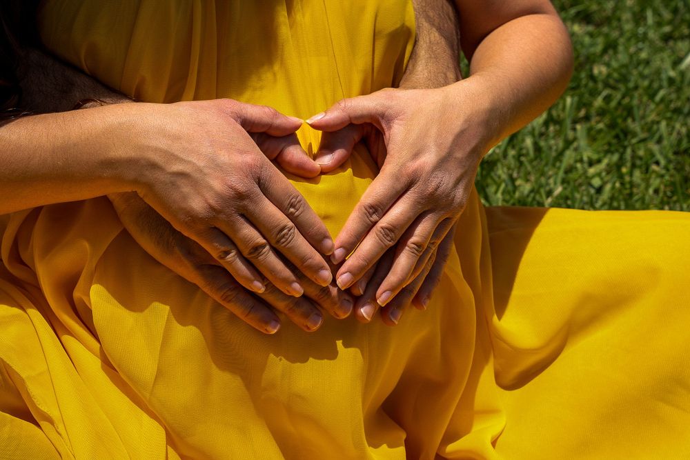 Close-up of hands forming a heart shape over a pregnant belly in a yellow dress