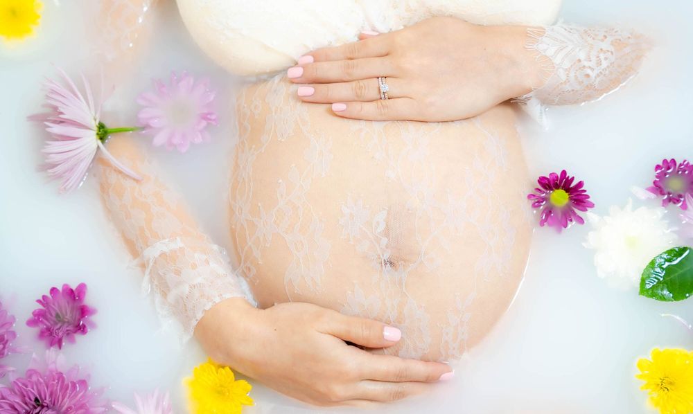 Close-up of a pregnant belly and hand resting on the edge of a flower-filled milk bath