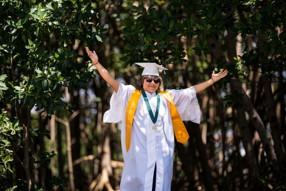 Graduate in cap and gown raising both arms in celebration surrounded by greenery