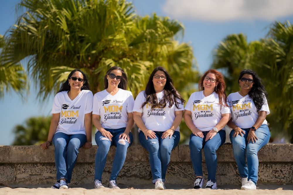 Group of graduates in matching t-shirts sitting together on a seawall with palm trees behind them