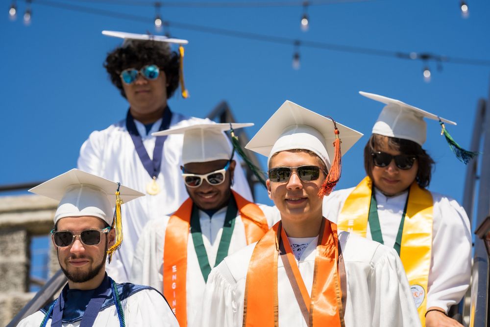 Small group of graduates in caps, gowns, and orange cords posing together under string lights