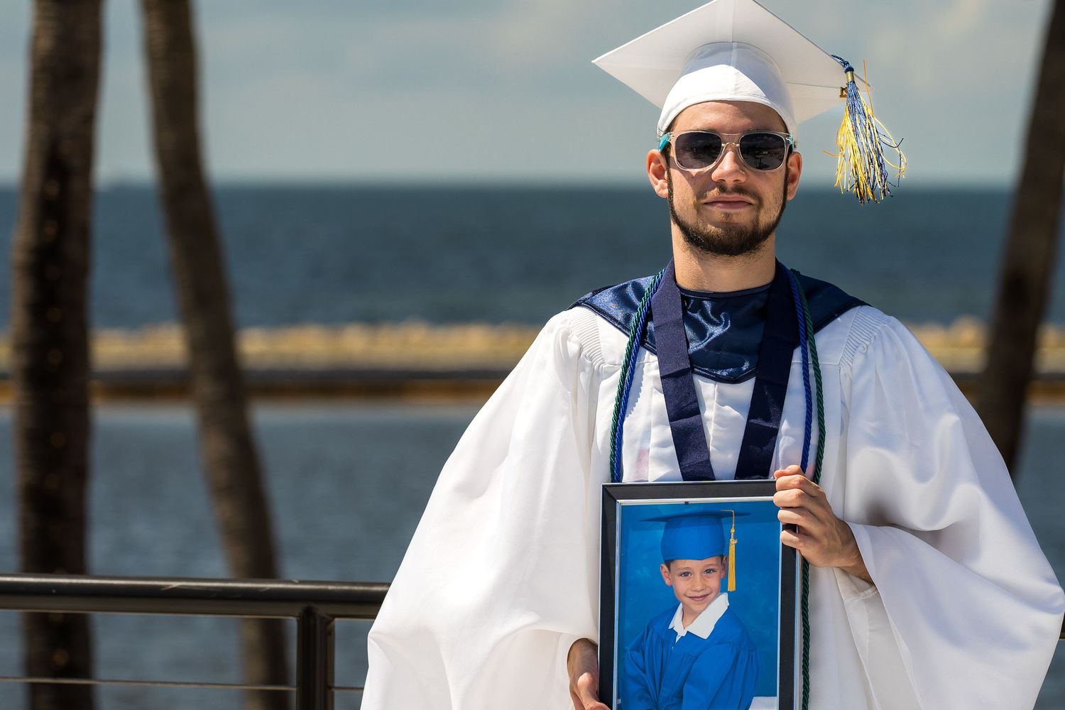 Graduate in cap and gown holding a framed photo while standing at a waterfront railing