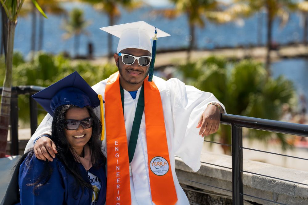 Graduate posing with a family member, both wearing graduation stoles, at a waterfront railing