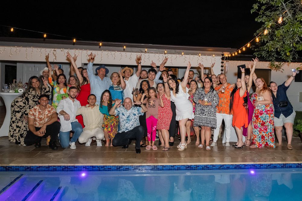 Large group of guests cheering together poolside at a nighttime celebration with string lights overhead