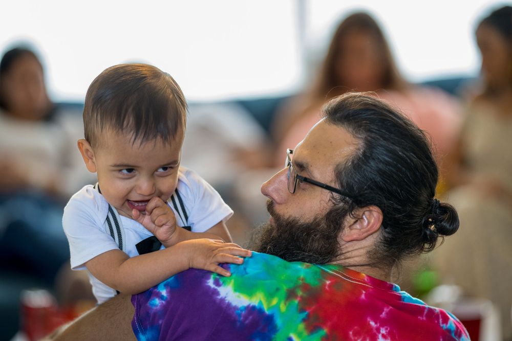 Father playing with his toddler, both wearing colorful tie-dye shirts, at an event