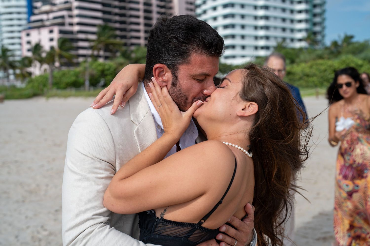 Couple kissing on the beach, the groom in a white suit, high-rise buildings behind them