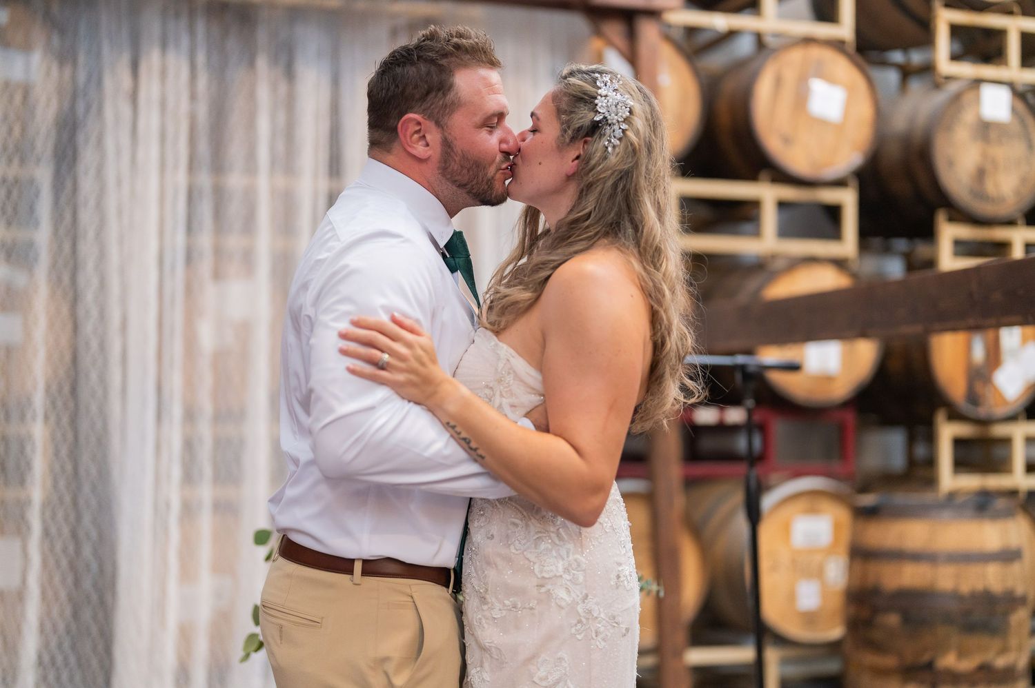 Groom and bride kissing in front of a wall of stacked barrels and white drapes