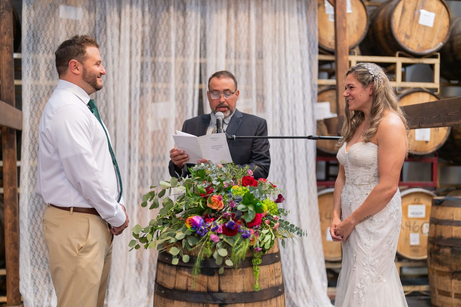 Officiant reading the ceremony while the bride and groom face each other, brewery barrels stacked behind them