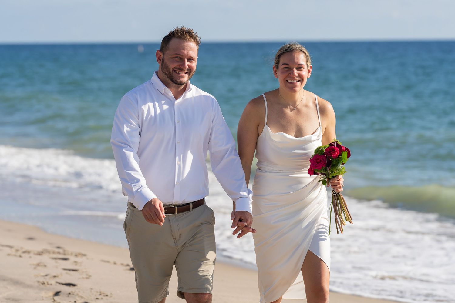Bride and groom walking hand in hand along the shoreline, the bride holding a red bouquet
