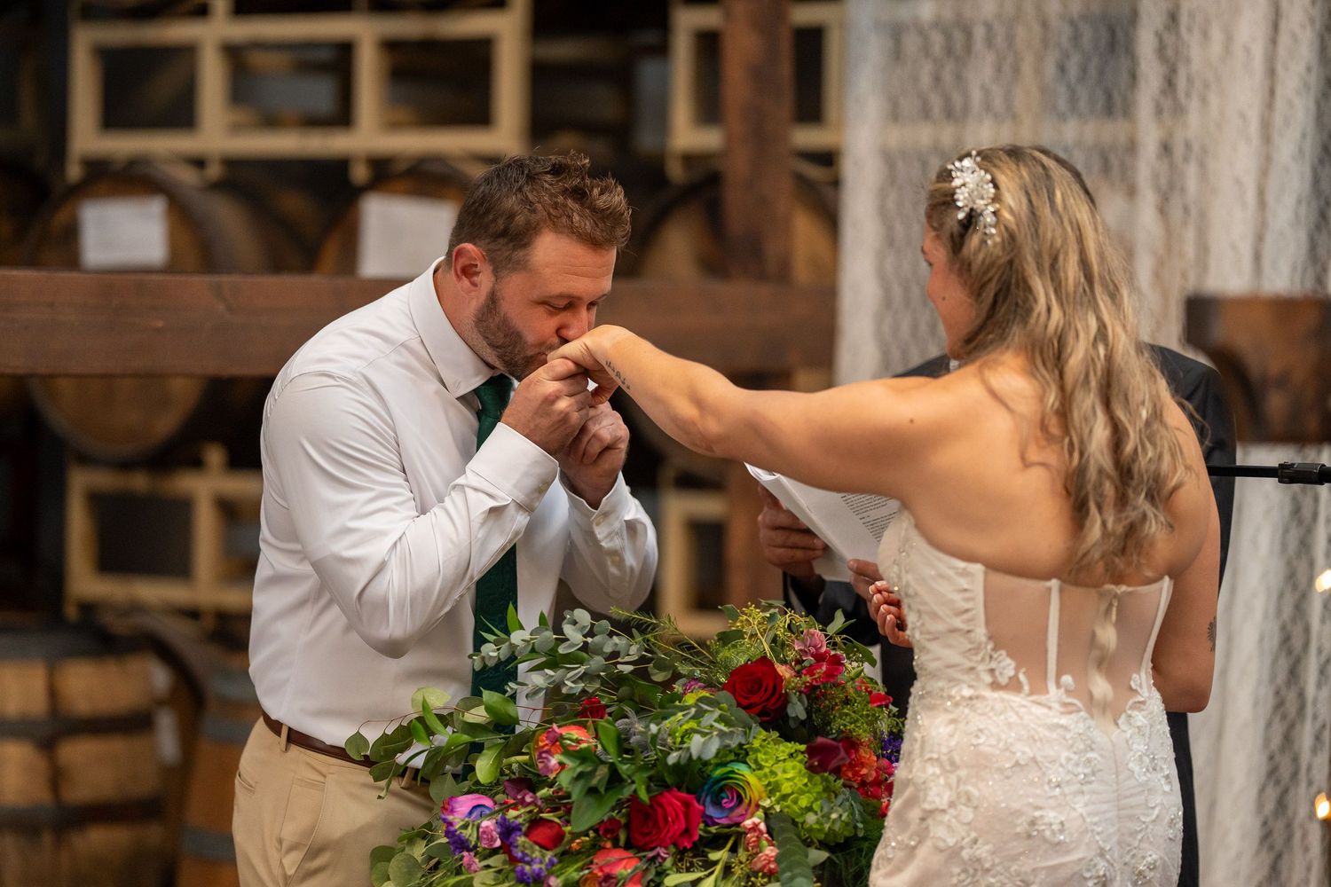 Groom kissing the bride's hand during the ceremony, a bouquet of red and yellow flowers in front