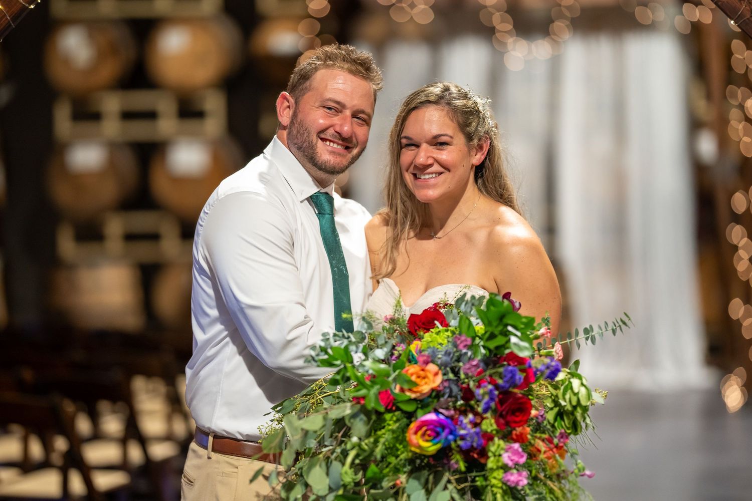 Smiling groom and bride holding a colorful bouquet inside a barrel room