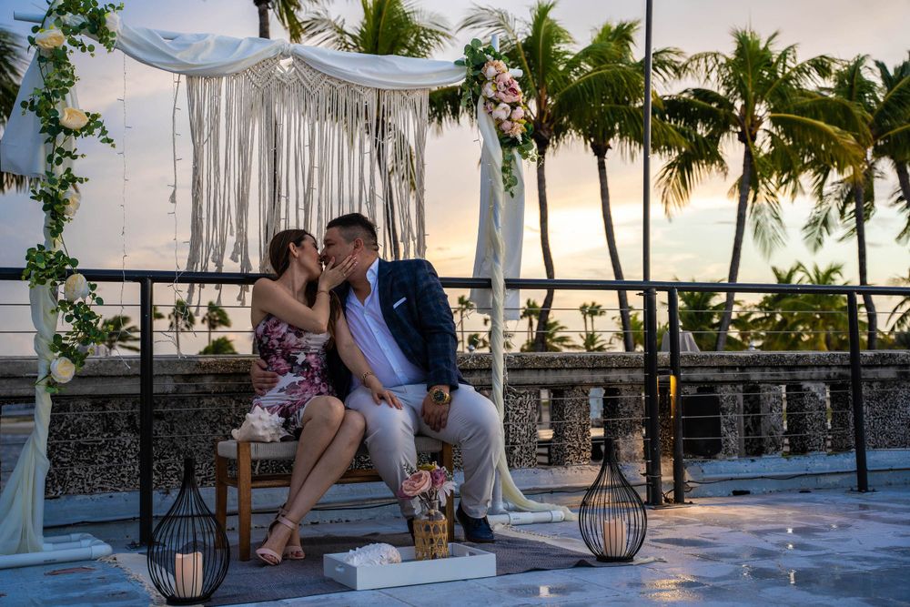 Bride and groom kissing under a macrame ceremony arch at sunset, palm trees along the water behind them