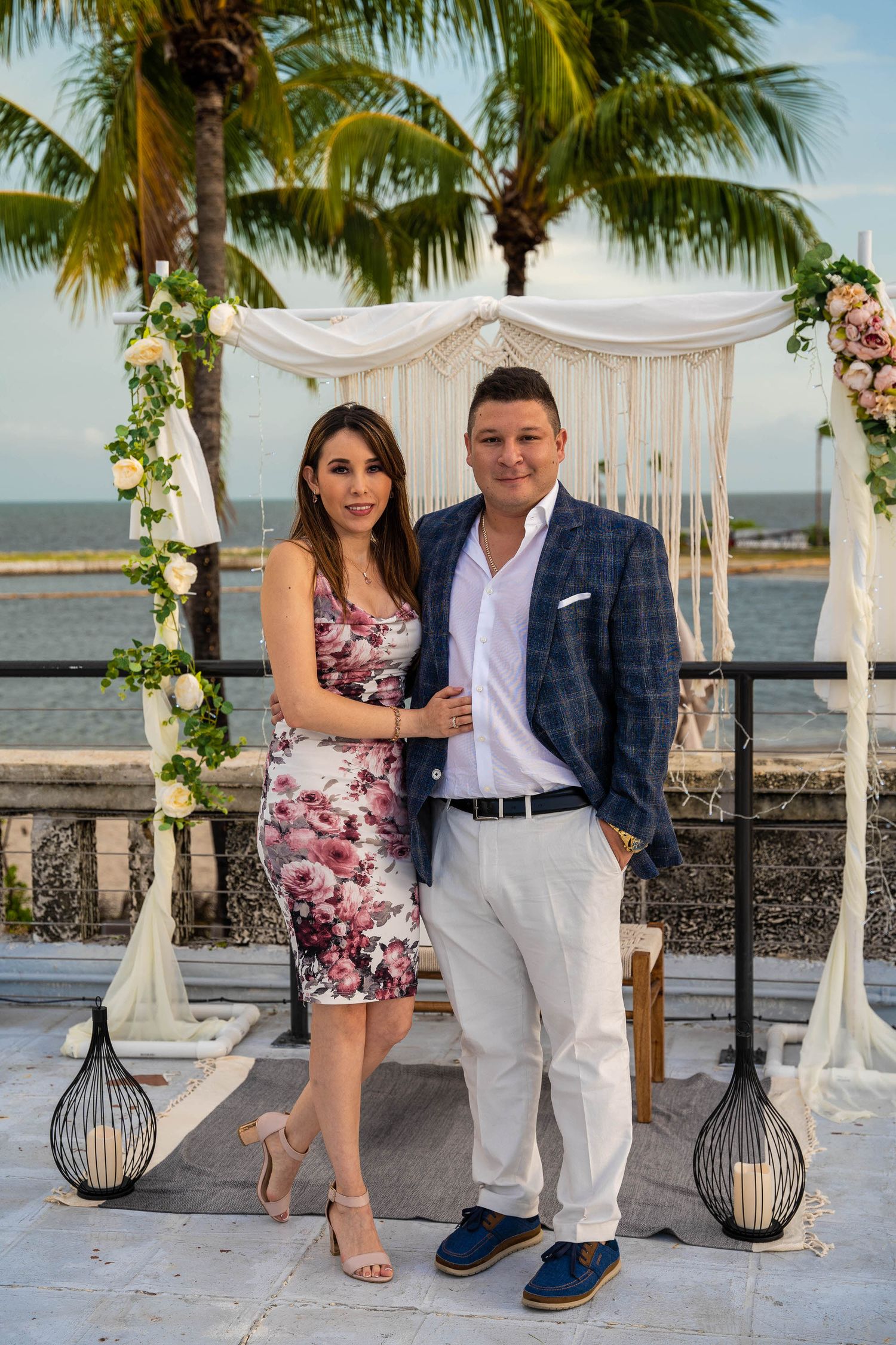 Bride and groom posed together under the floral ceremony arch, palm trees and water in the background