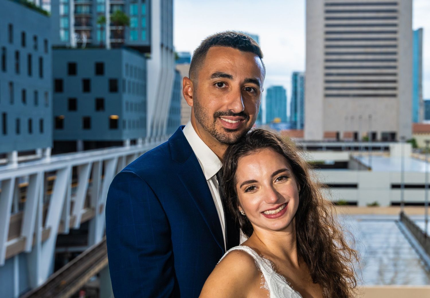 Groom in a navy suit leaning in beside the smiling bride with a city skyline behind them