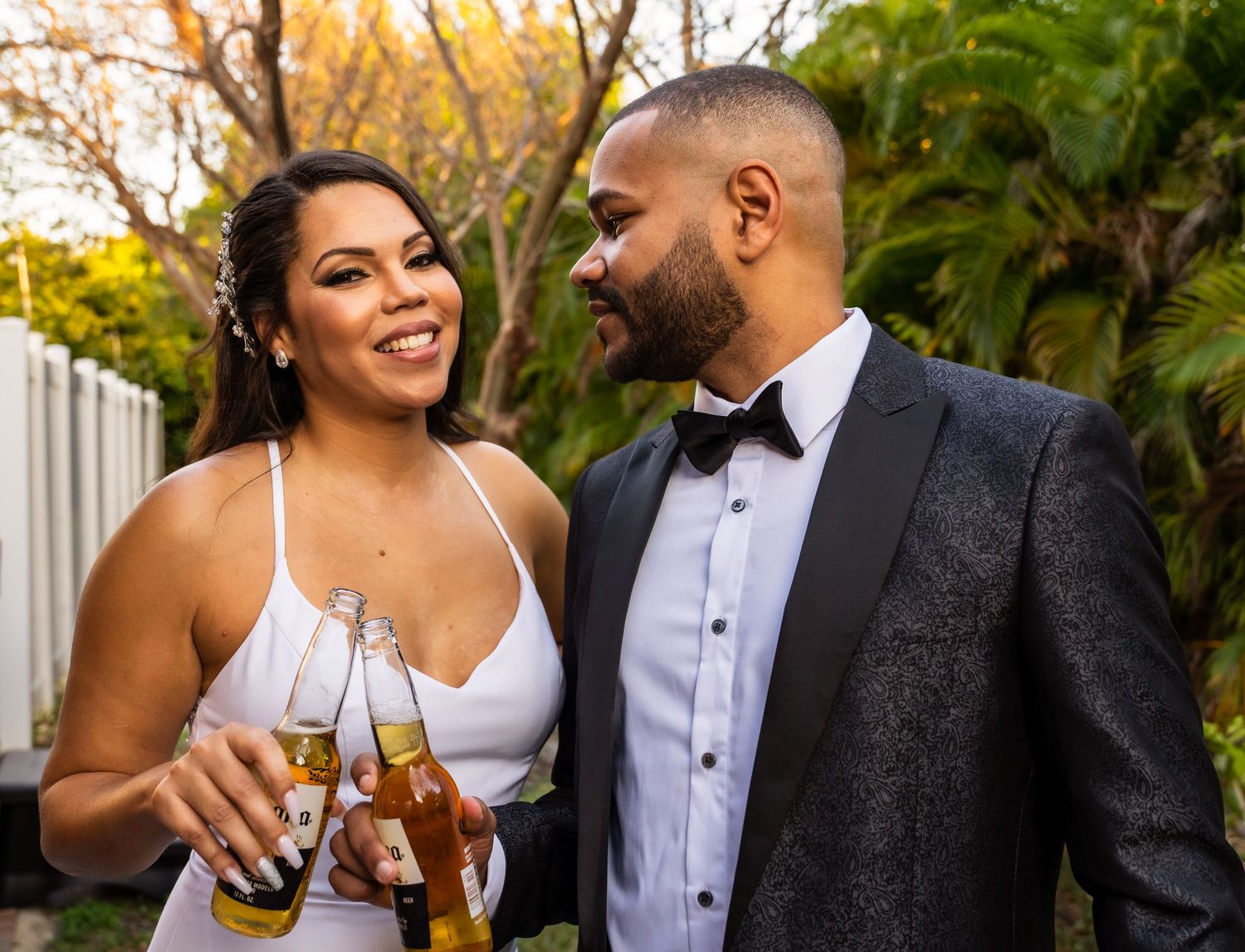 Bride and groom clinking beer bottles, smiling at each other among palm leaves