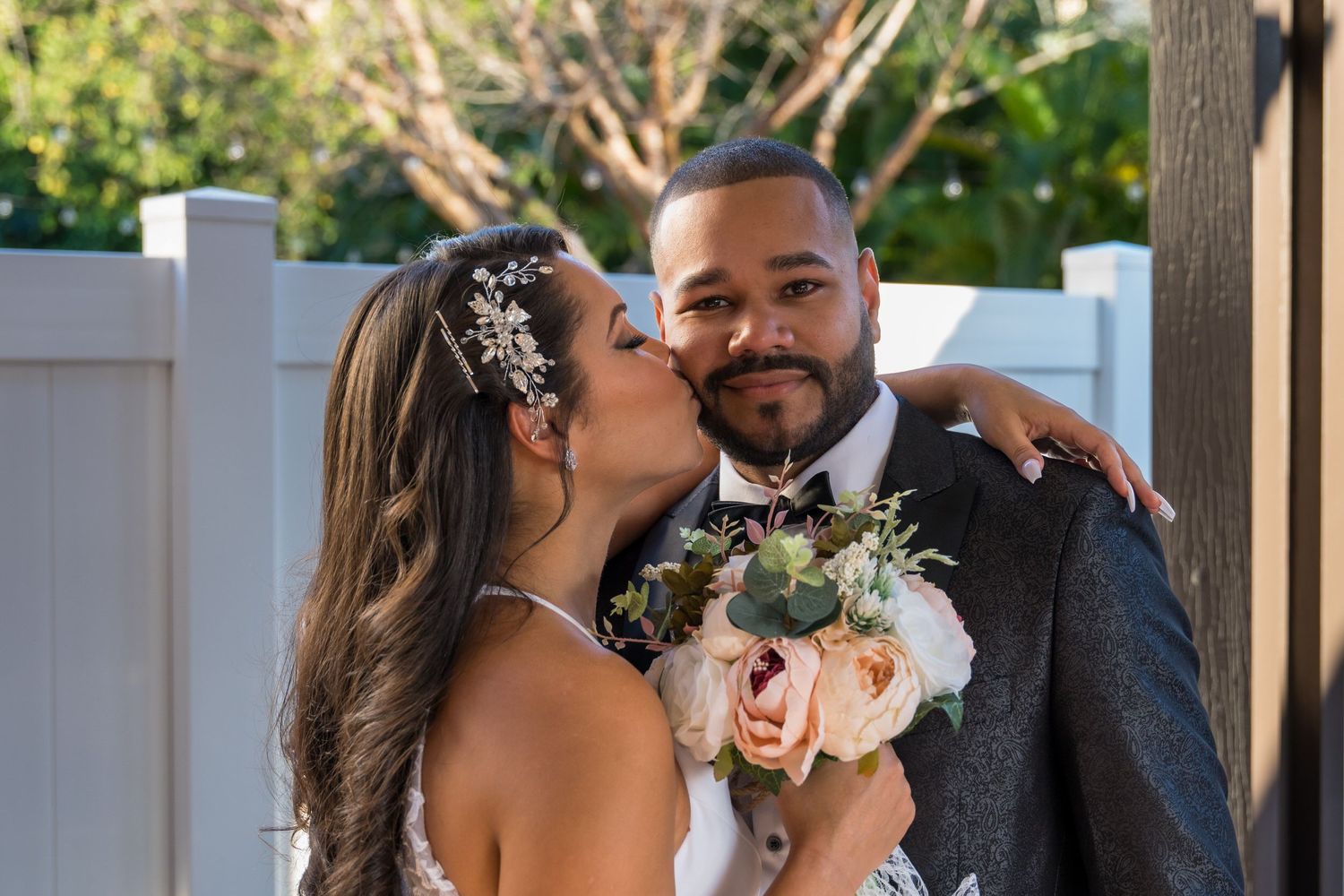 Bride kissing the groom on the cheek, holding a pastel bouquet, white fence behind