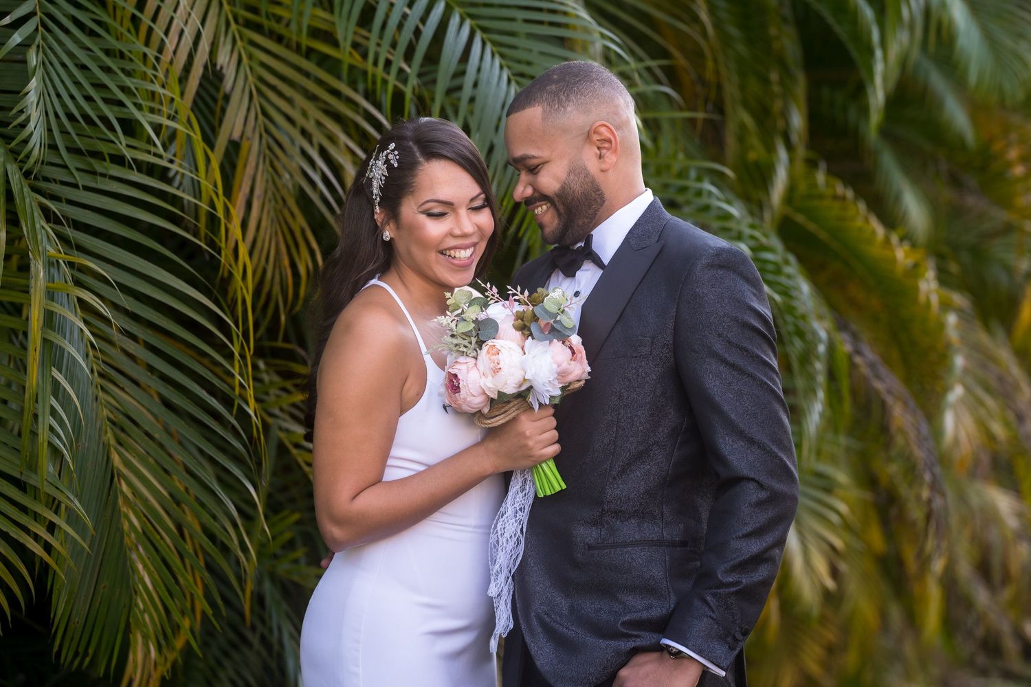 Bride and groom laughing together in front of palm fronds, the bride holding a bouquet