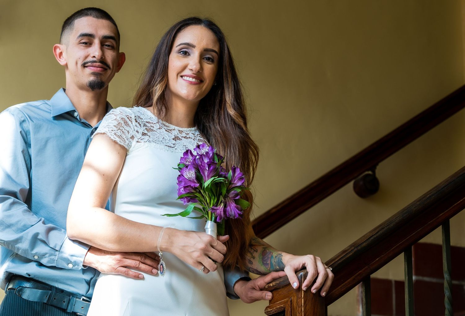 Bride with a purple bouquet and groom smiling together beside a staircase