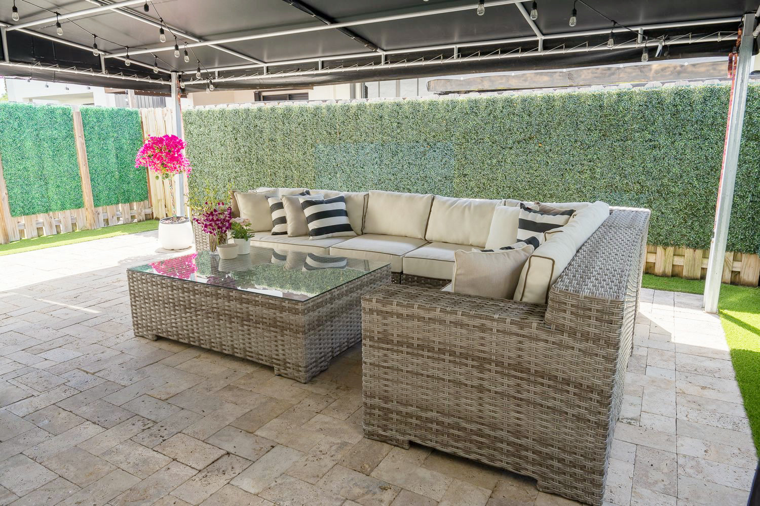 Covered patio lounge with a wicker sectional and string lights against a green hedge wall