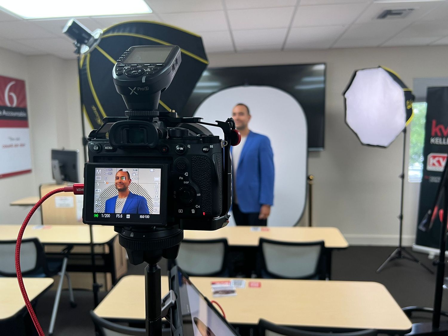 Cinema camera on a tripod with a softbox light, framing a subject in a blue blazer against a portable backdrop