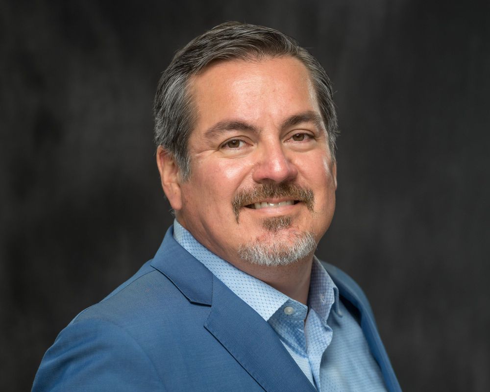 Studio headshot on a dark backdrop, light blue shirt and blazer