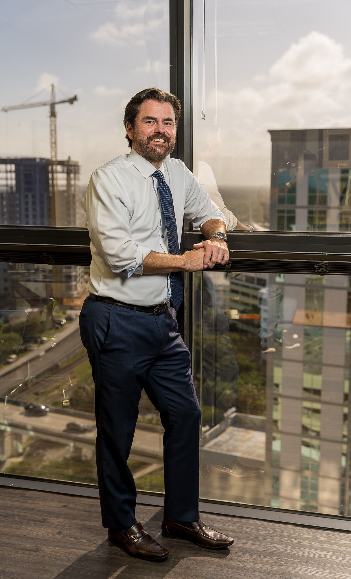 Full-length portrait of a man in a tie and dress shirt leaning on a glass railing above the city