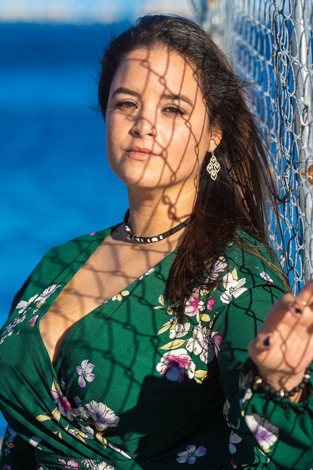 Model in a dark floral top posing behind a chain-link fence at dusk