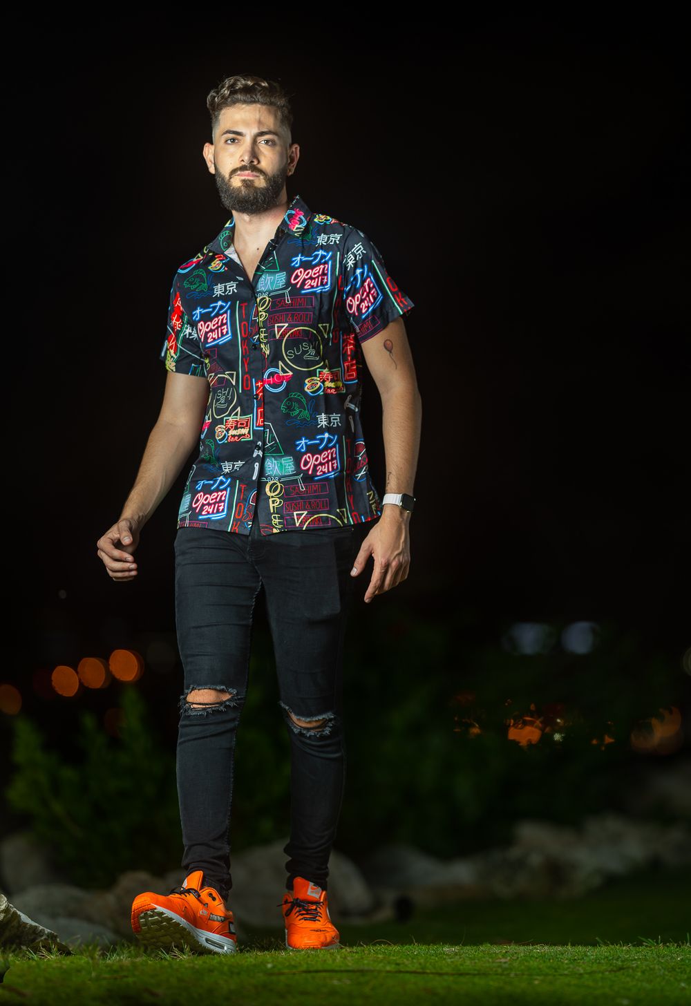 Male model in a bright floral shirt and ripped black jeans standing against a dark night backdrop