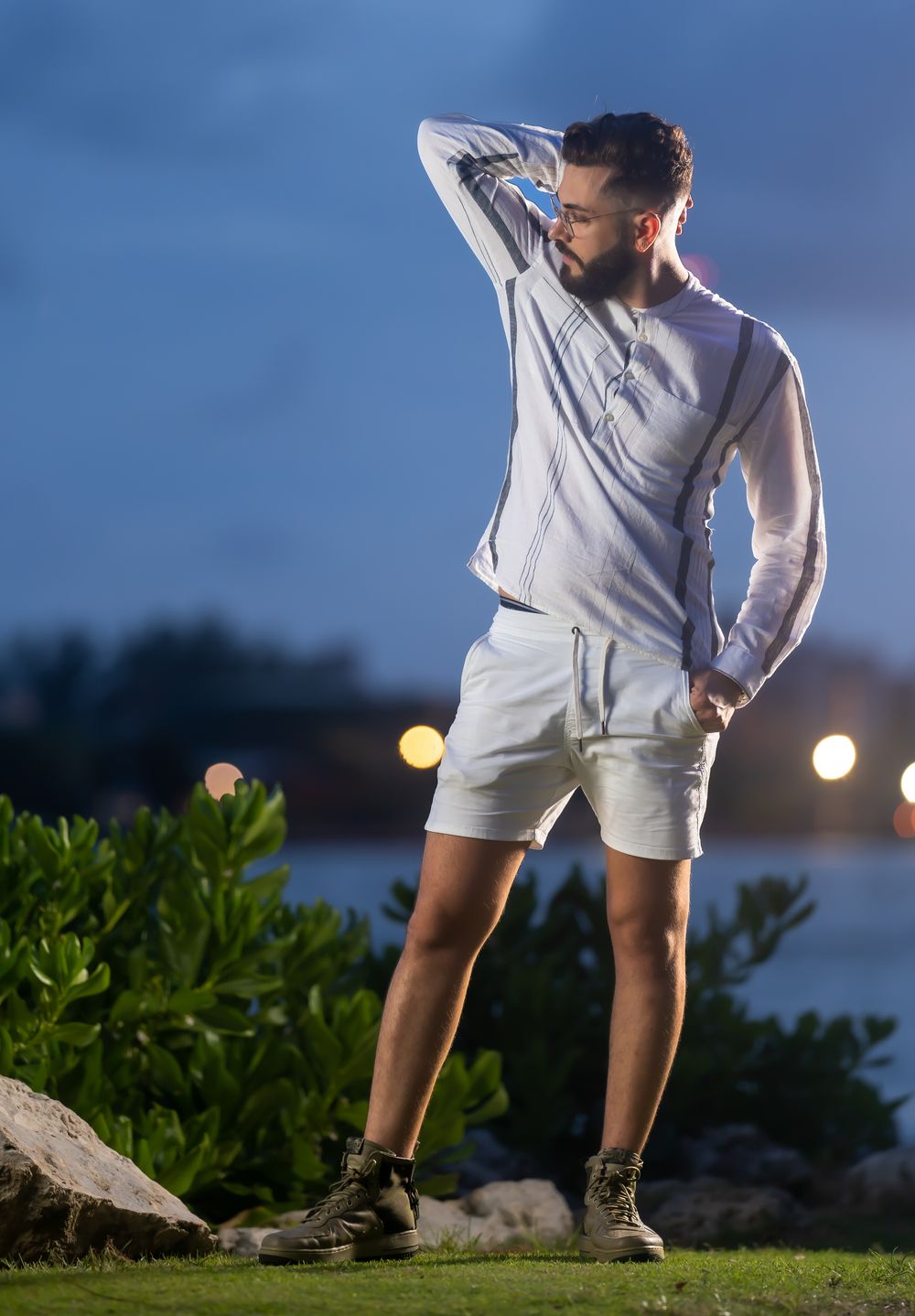 Male model in a white shirt and shorts striking a pose on the grass at dusk with city lights behind him