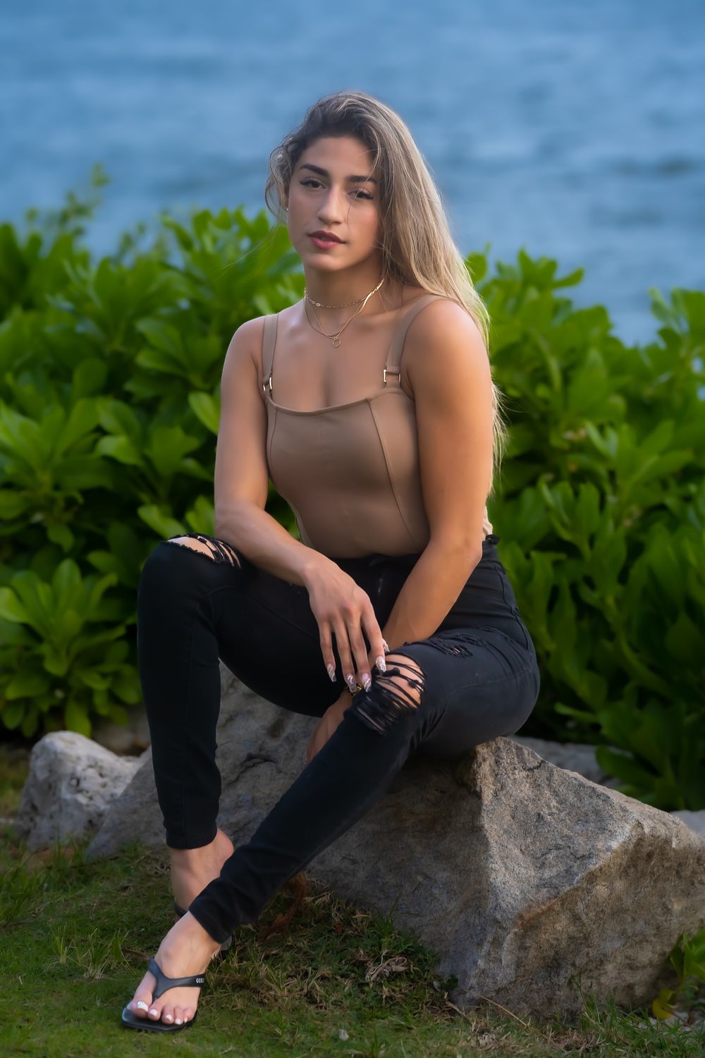 Model in a brown top and black pants seated on a rock with greenery and water behind her at dusk