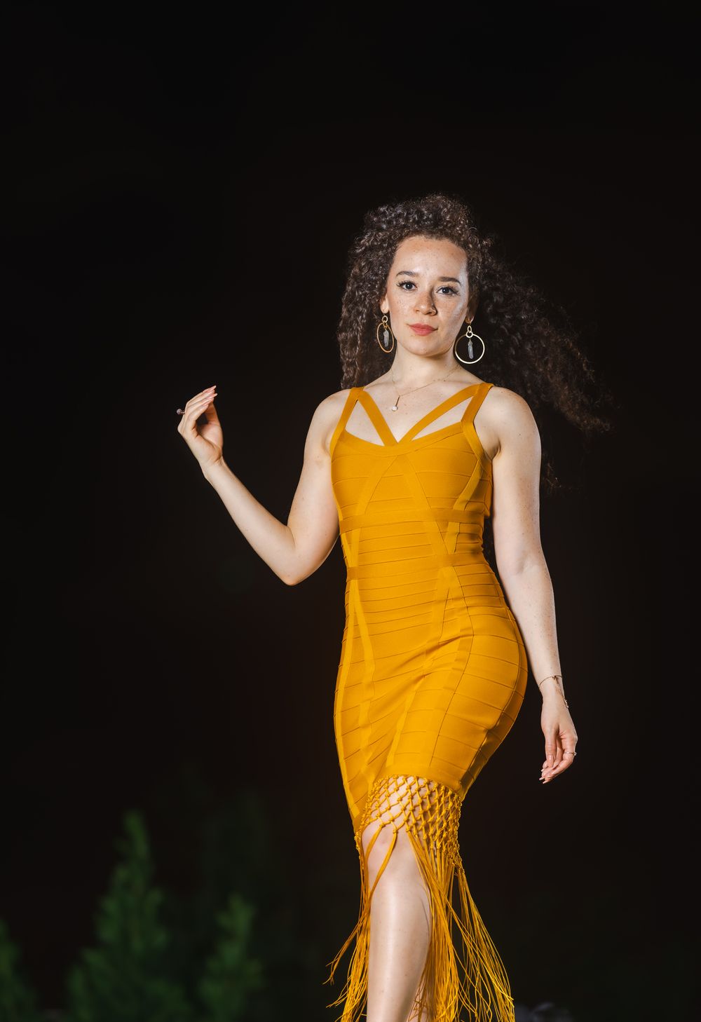 Model in a mustard fringe dress mid-pose with one arm out against a dark night backdrop