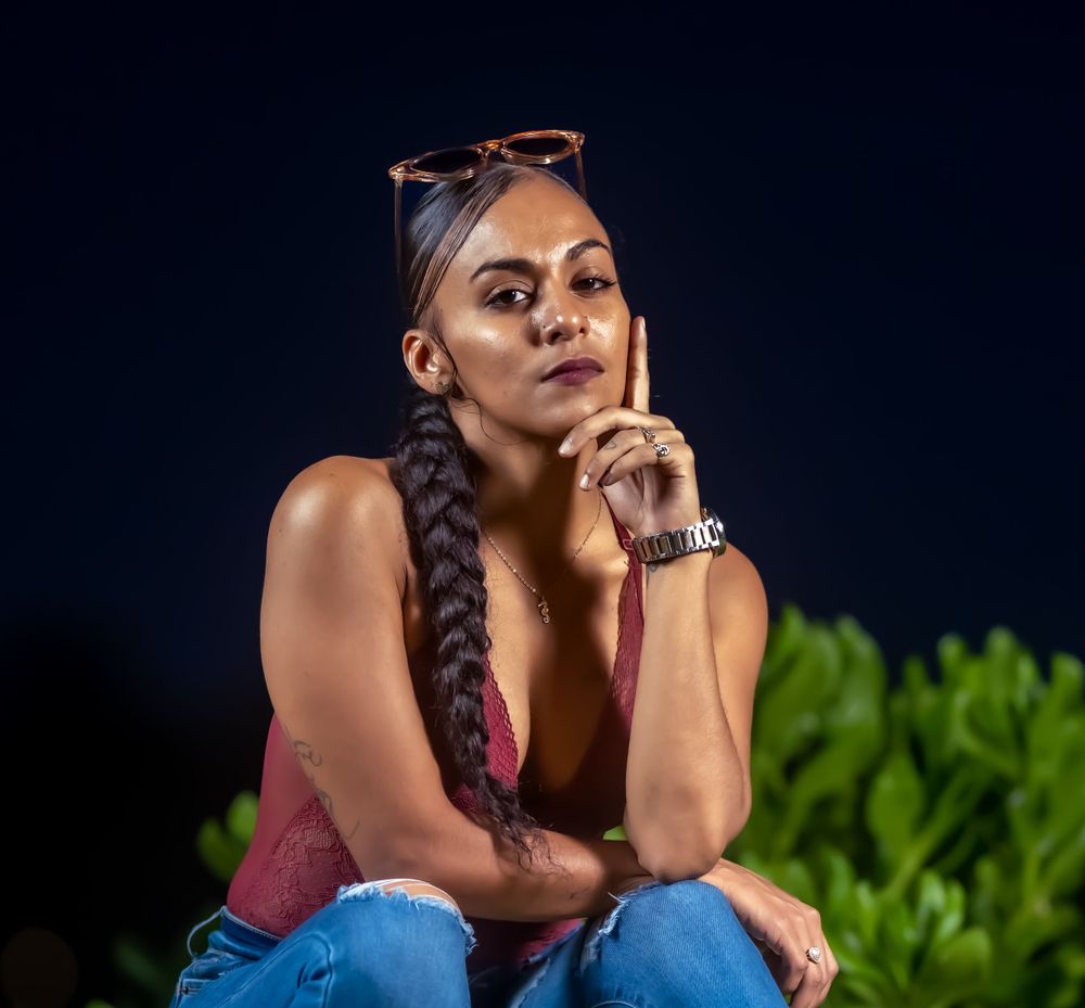 Model in a burgundy top posing with her hand near her chin against greenery at night