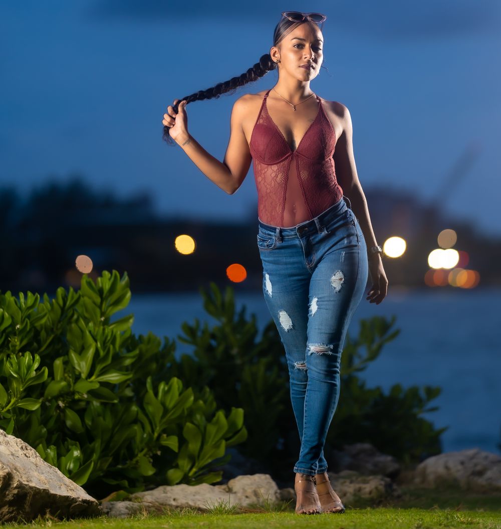Model in a burgundy top and ripped jeans holding her braid with city lights glowing behind her at dusk